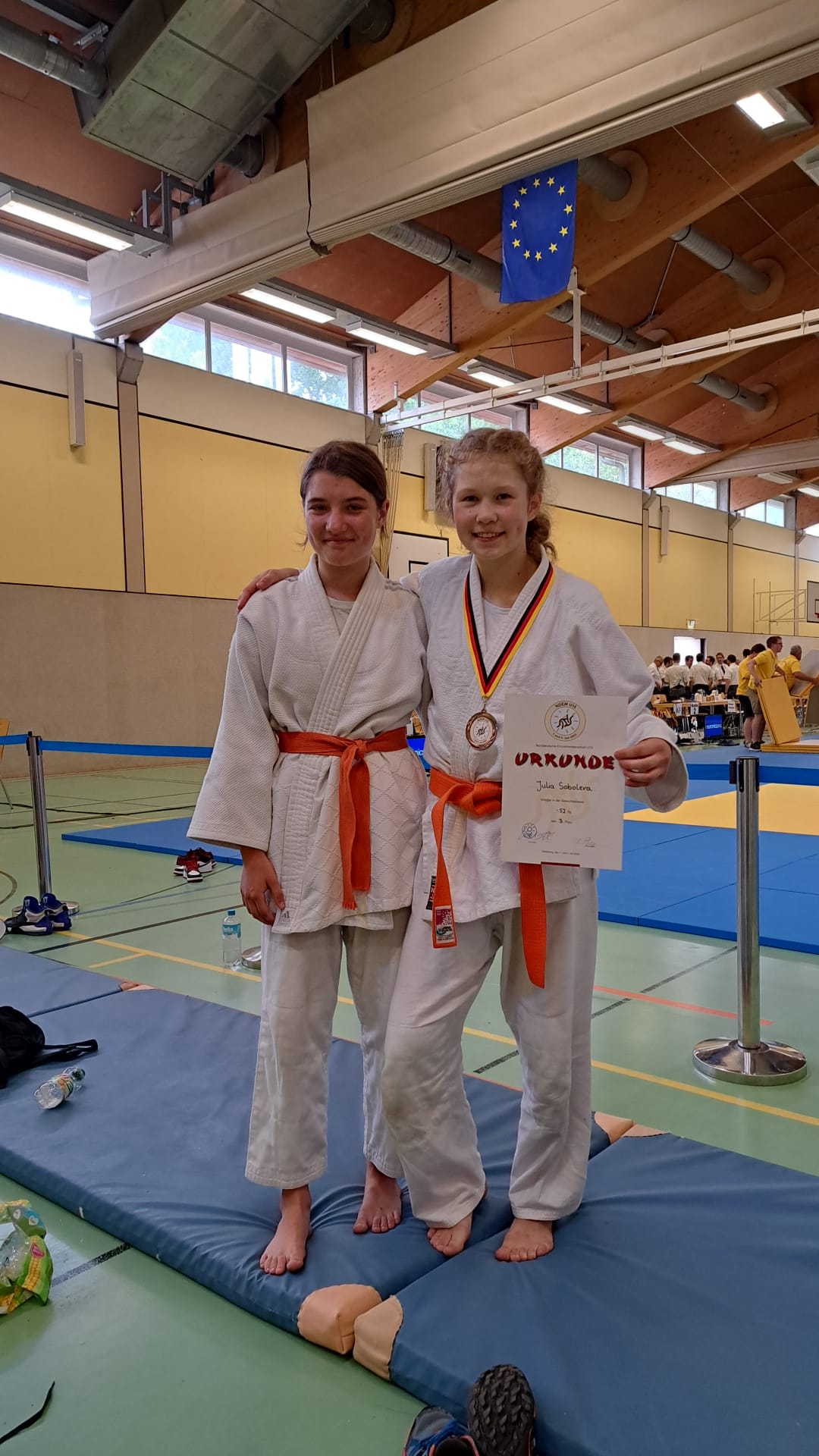 Zwei Judo-Kämpferinnen in weißen Anzügen mit orangen Gürteln stehen auf einer Matte und halten eine Urkunde.