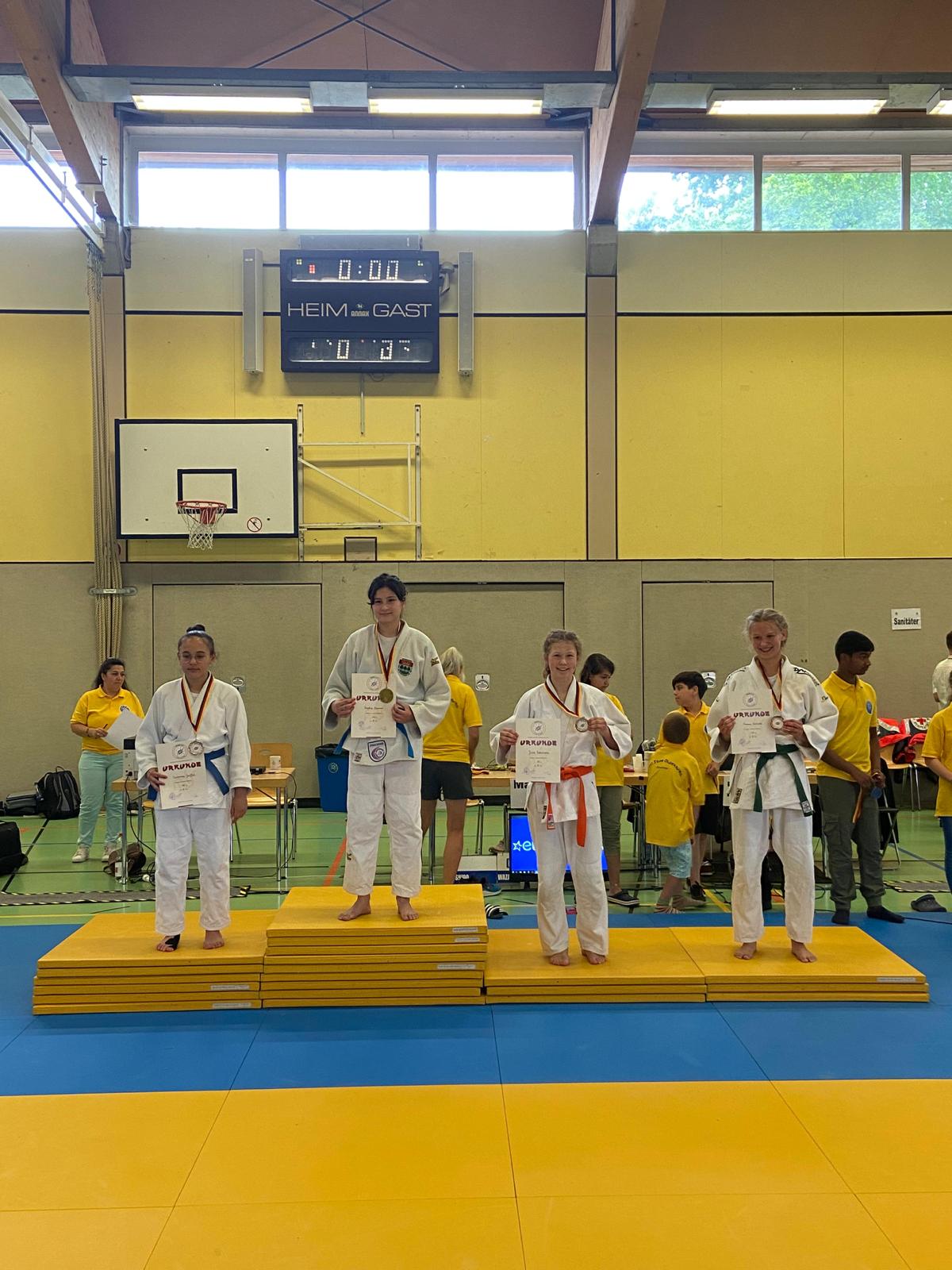 Vier Judokas stehen auf einem Siegerehrungspodest, halten Medaillen und Urkunden in der Sporthalle.