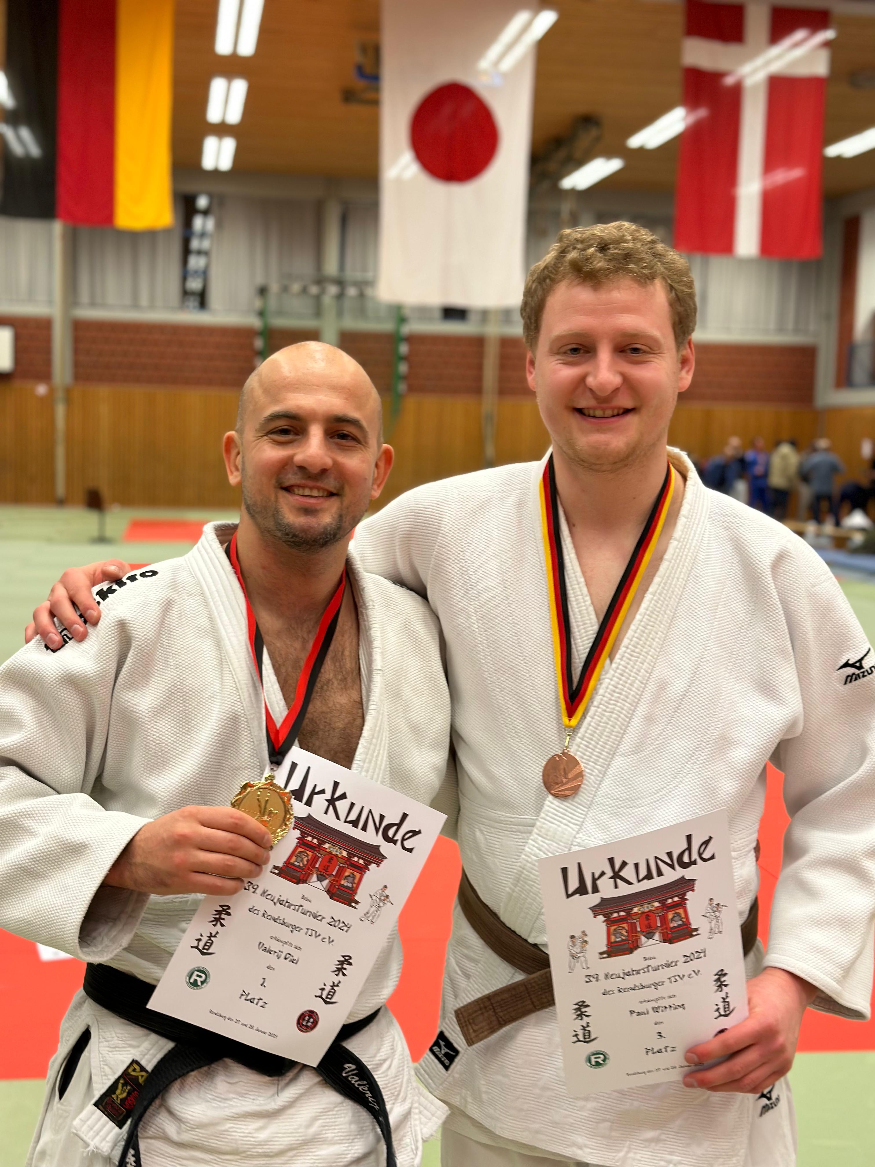 Zwei Judoka in weißen Anzügen halten Urkunden und Medaillen vor einer Sporthalle mit Flaggen im Hintergrund.