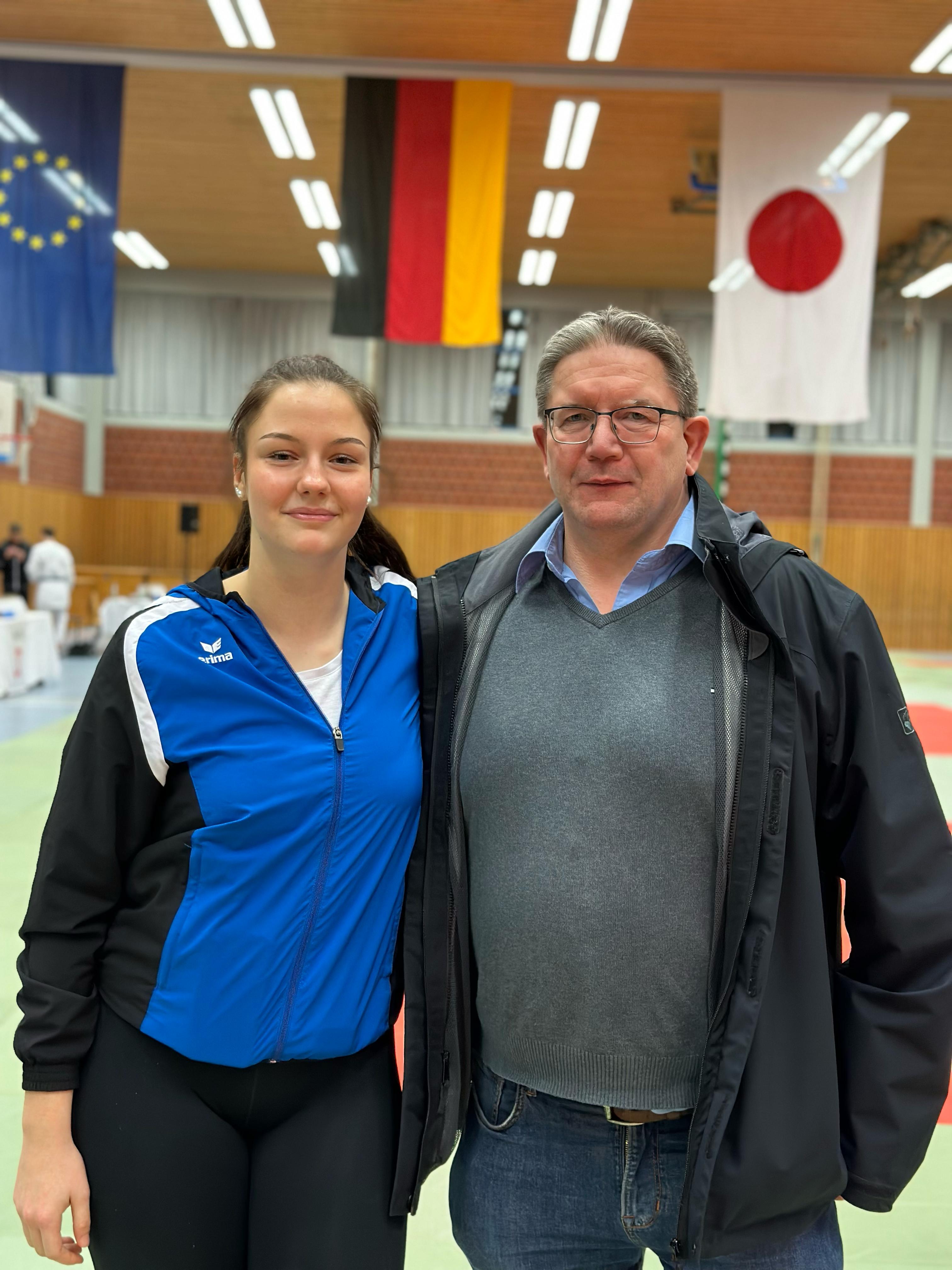 Junge Sportlerin mit blauer Jacke steht neben einem Mann in einem Trainingsraum mit Fahnen im Hintergrund.