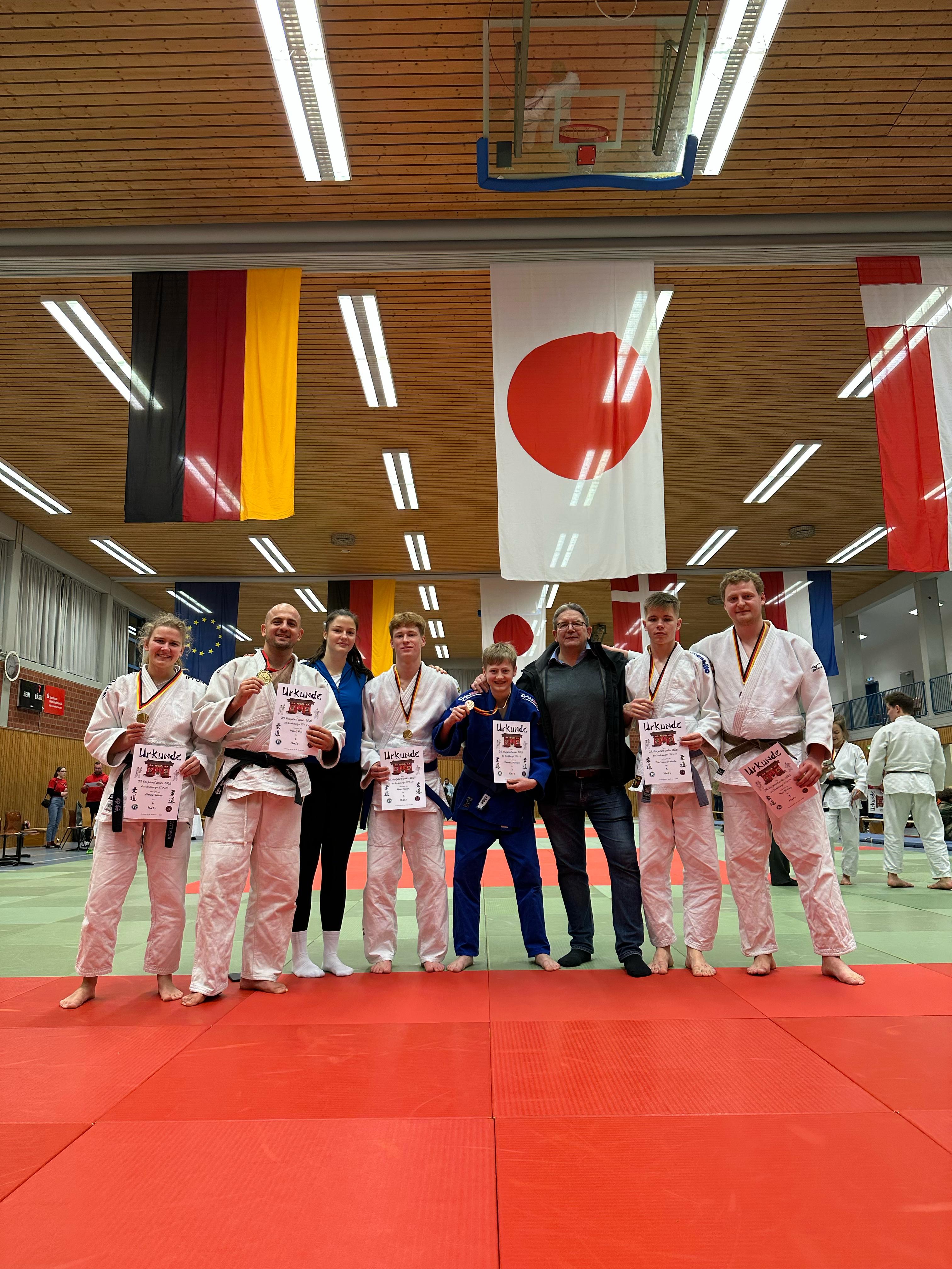 Gruppe von Judoka in weißen und blauen Anzügen mit Urkunden auf einer roten Matte, umgeben von Flaggen im Hintergrund.