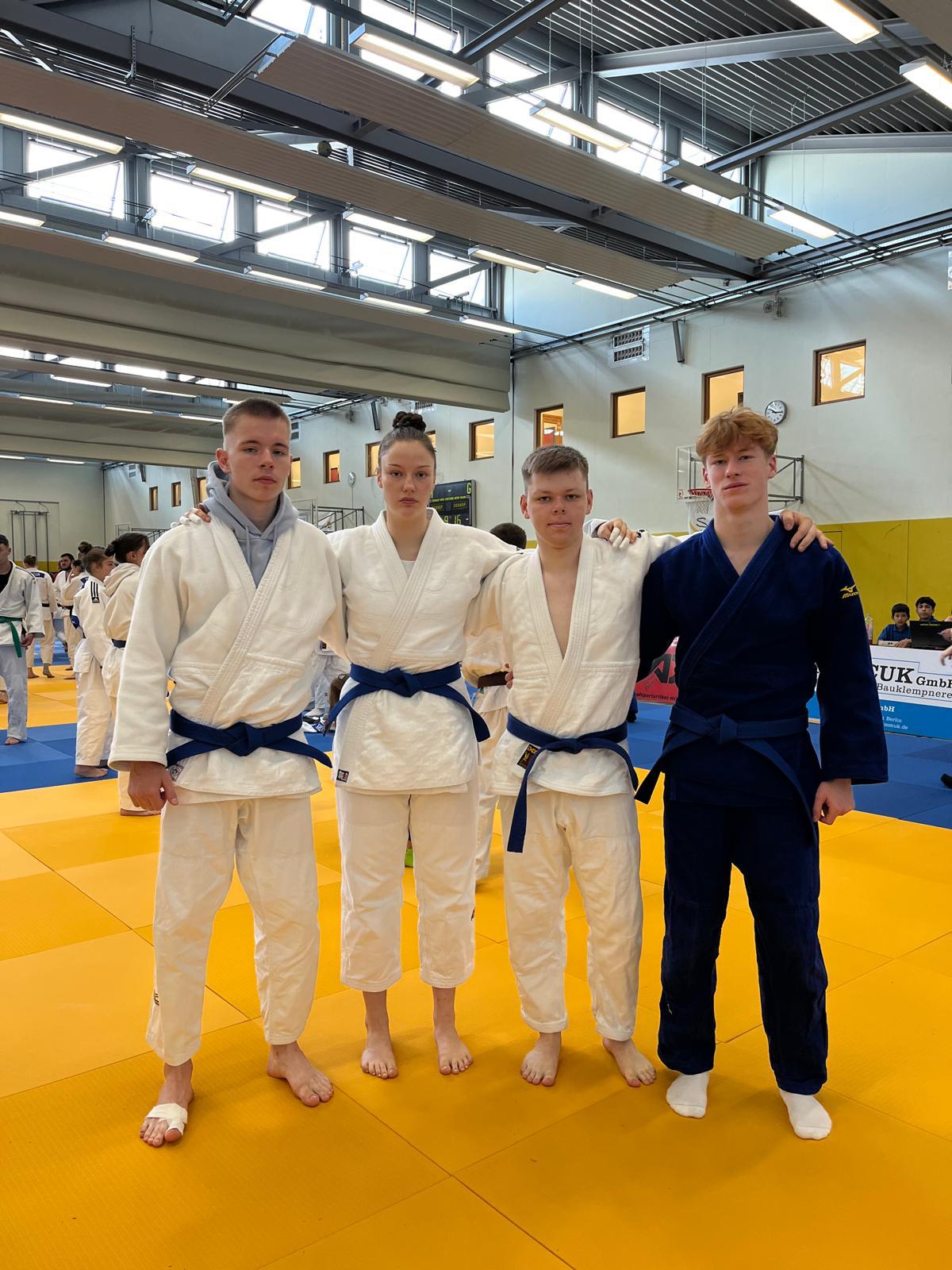 Vier Judoka in Trainingsanzügen mit blauen Gürteln posieren auf einer Judomatte in einer Halle.