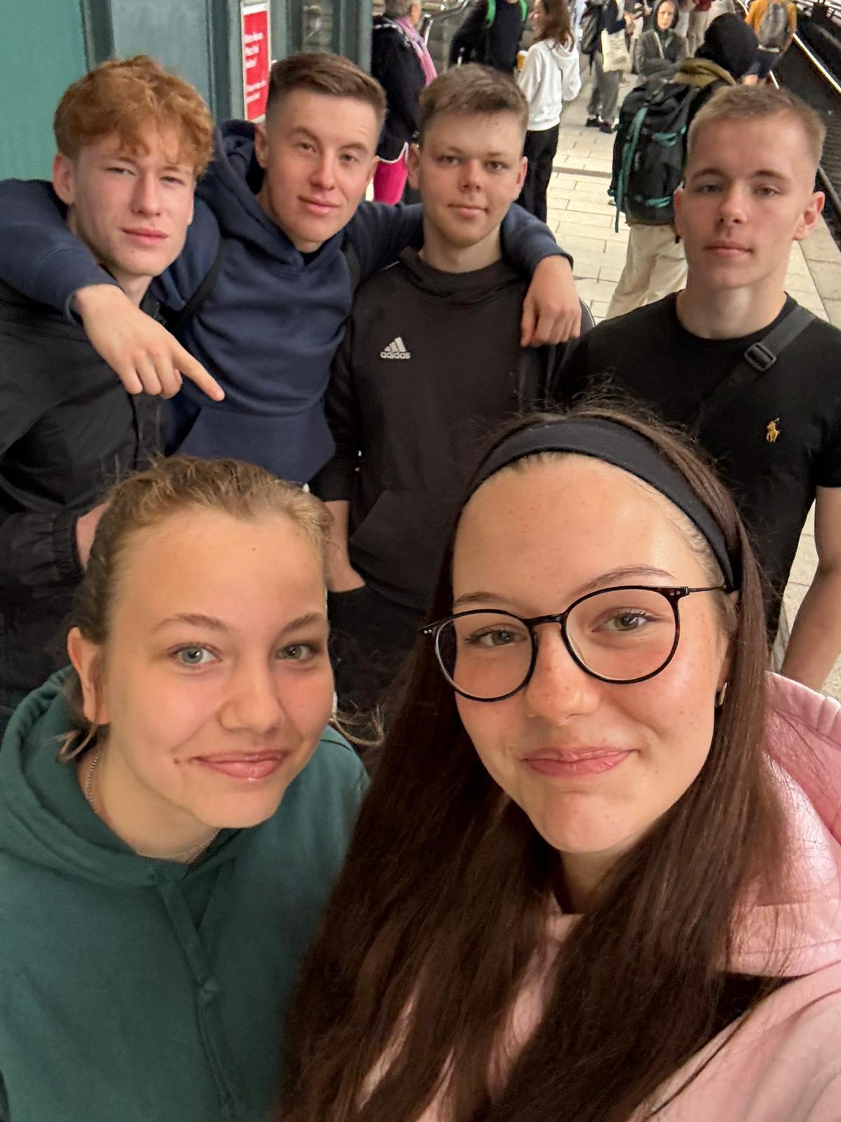 Gruppe junger Menschen posiert lächelnd in einer U-Bahn-Station, im Hintergrund sind weitere Fahrgäste sichtbar.