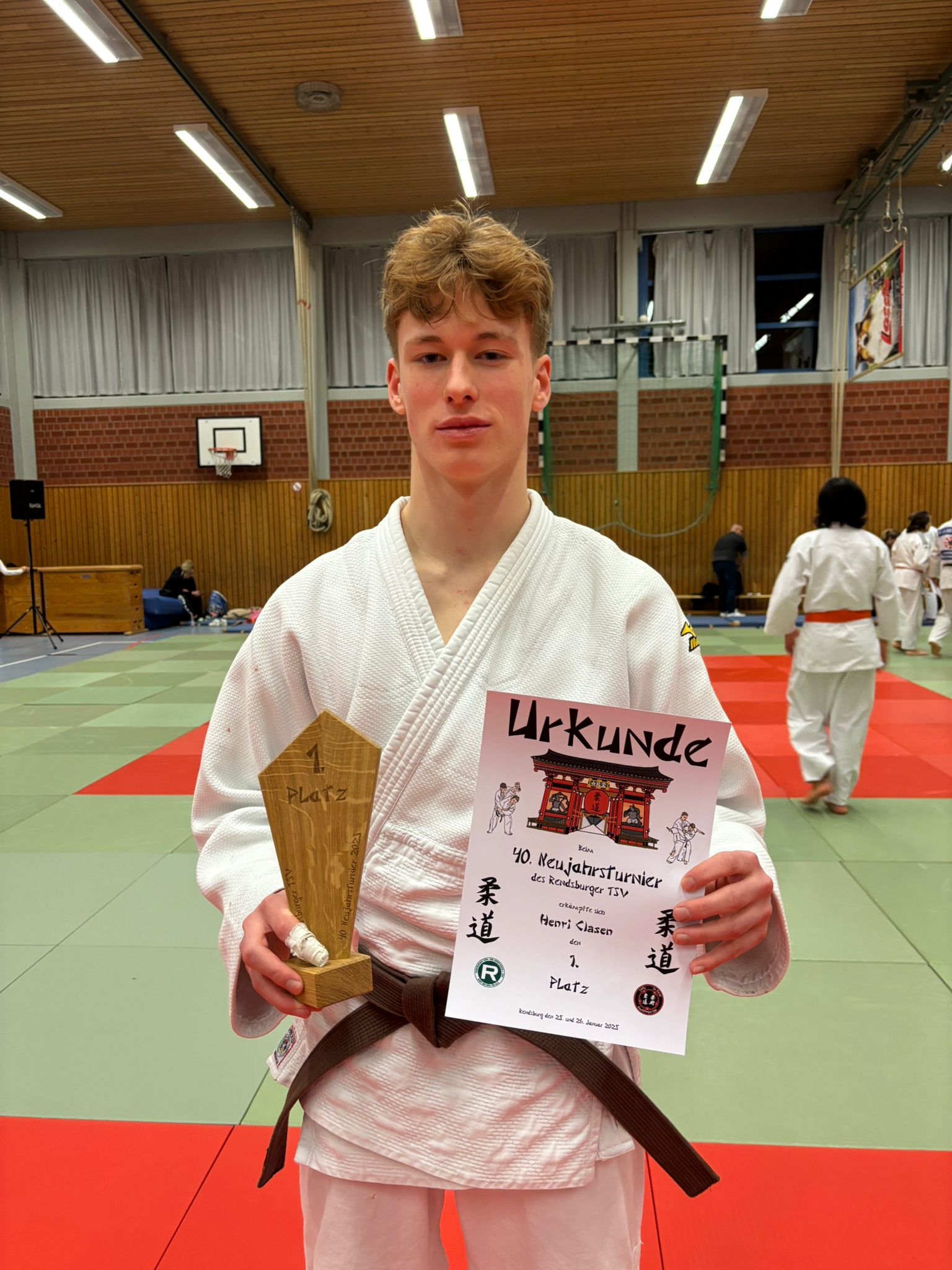 Junge im Judoanzug hält einen ersten Platz Pokal und eine Urkunde in einer Turnhalle mit weiteren Judokämpfern.