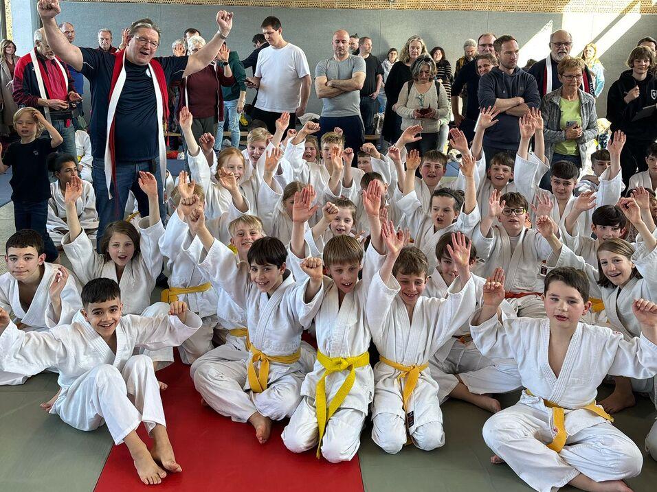 Gruppenselfie von Kindern in weißen Judojacken mit gelben Gürteln, umgeben von Zuschauern in einer Sporthalle.