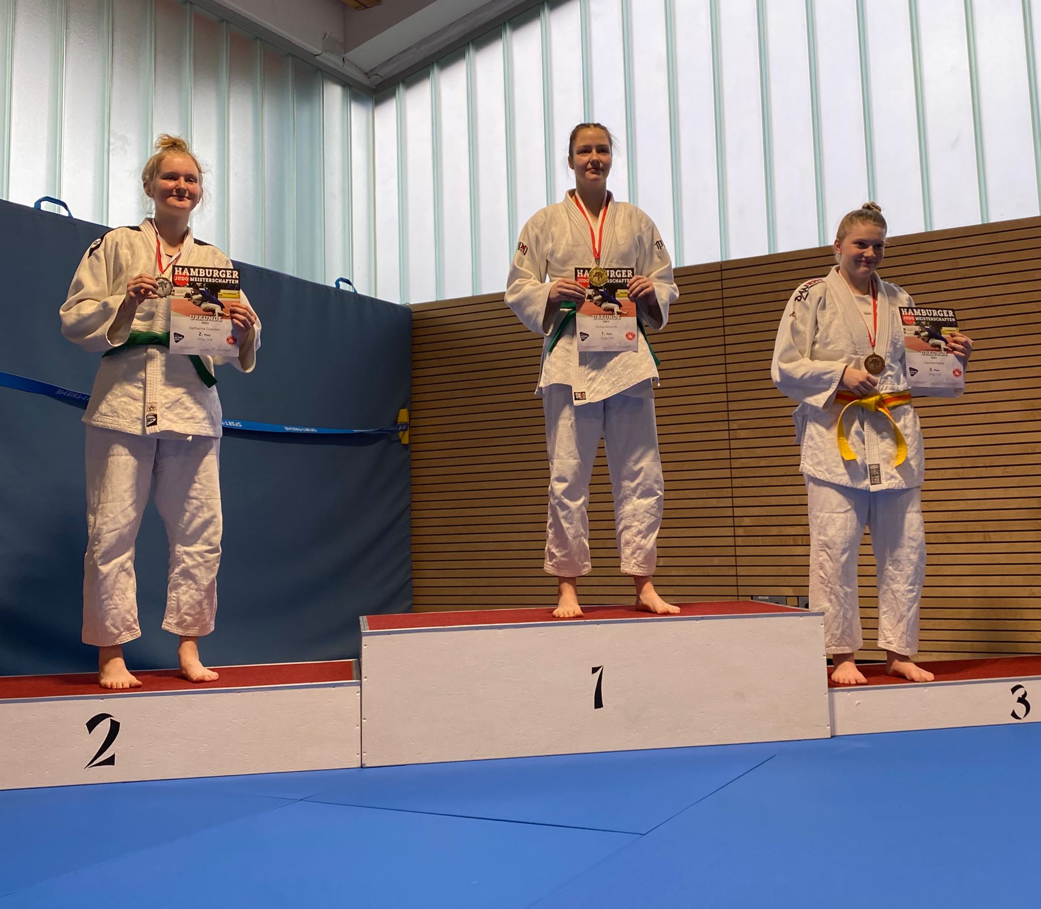 Drei Judokas stehen auf dem Siegertreppchen, halten Urkunden und tragen Medaillen in einer Sporthalle.