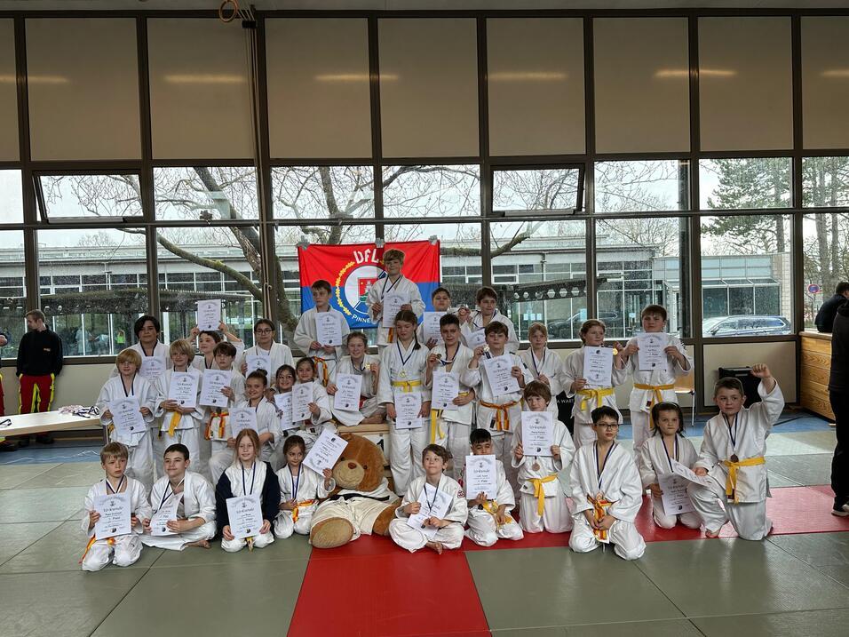 Gruppe von Kindern in Karateanzügen mit Urkunden, umgeben von Fenstern und einer rot-blauen Flagge im Hintergrund.