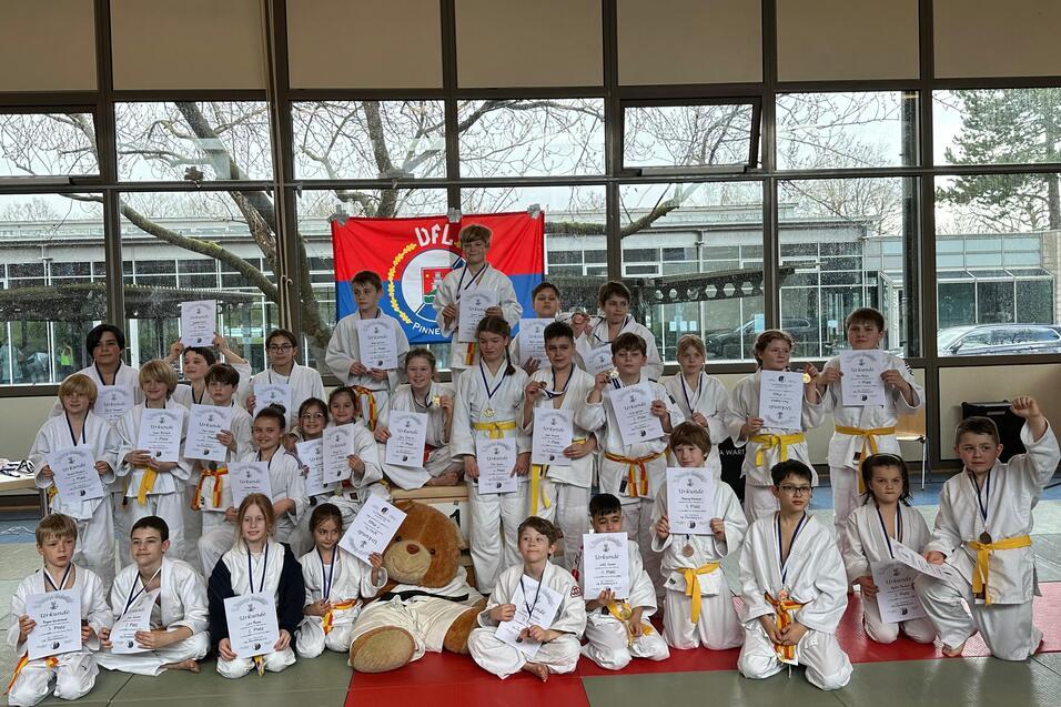 Gruppe von Kindern in Karateanz&uuml;gen mit Urkunden, umgeben von Fenstern und einer rot-blauen Flagge im Hintergrund.