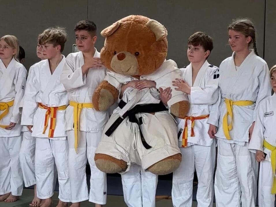 Gruppe von Judokas in weißen Anzügen mit gelben Gürteln und einem großen Teddybären im Judogi auf einer Matte.
