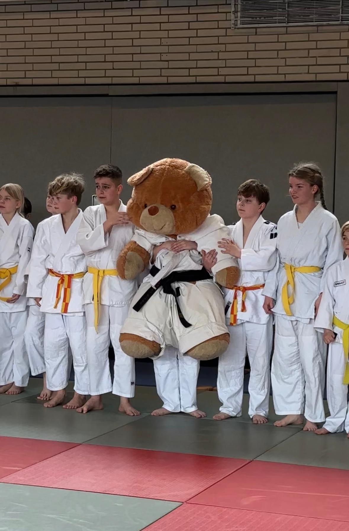 Gruppe von Judokas in weißen Anzügen mit gelben Gürteln und einem großen Teddybären im Judogi auf einer Matte.