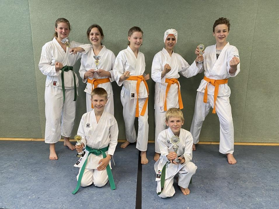 Sieben Karateka in Weiß mit verschiedenen Gürtelfarben zeigen Pokale, posieren stolz vor einer Wand.