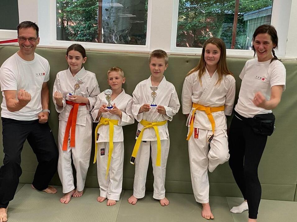 Gruppe von fünf Karateka in Weiß, mit Trophäen, lächelnd auf einer Matte in einem Trainingsraum.