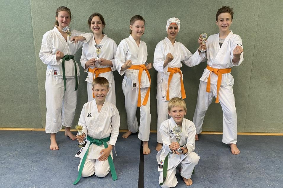 Sieben Karateka in Wei&szlig; mit verschiedenen G&uuml;rtelfarben zeigen Pokale, posieren stolz vor einer Wand.