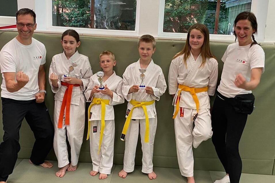 Gruppe von f&uuml;nf Karateka in Wei&szlig;, mit Troph&auml;en, l&auml;chelnd auf einer Matte in einem Trainingsraum.