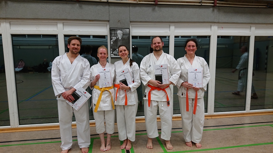 F&uuml;nf Karateka in wei&szlig;en Gi mit Urkunden stehen l&auml;chelnd vor einem Fenster in einem Dojo.