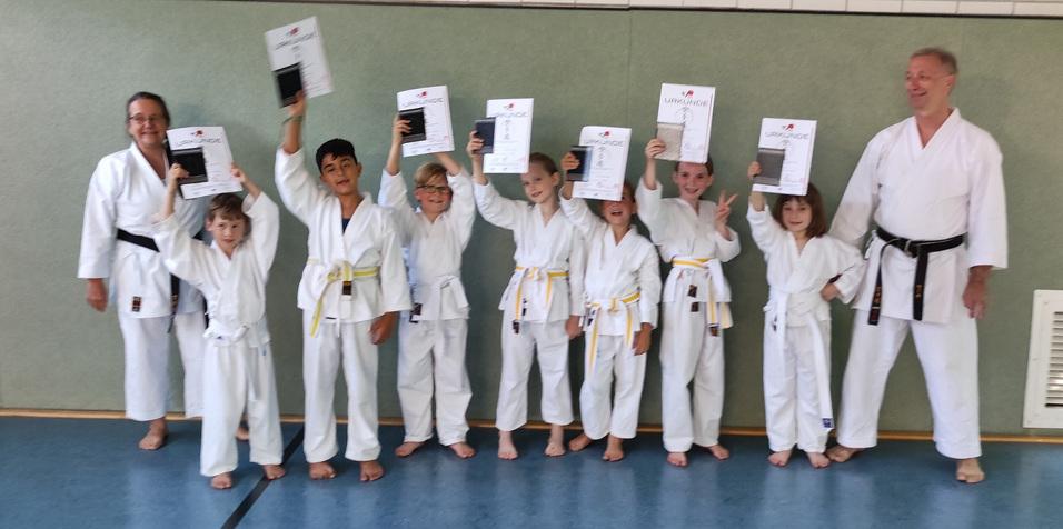 Gruppe junger Karateka in Gi hält stolz Urkunden in einem Trainingsraum. Ein Trainer steht lächelnd daneben.