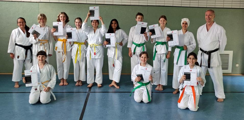 Gruppenszene von Karateka in weißen Gi, die stolz ihre Urkunden in der Sporthalle halten. Farbige Gürtel sichtbar.