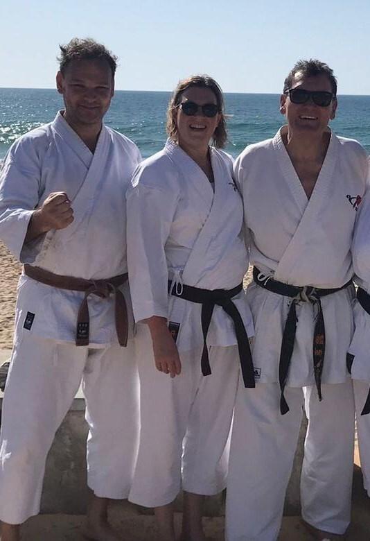 Drei Karateka in weißen Gi mit Braungurt und zwei Schwarzgurten posieren lächelnd am Strand vor dem Meer.