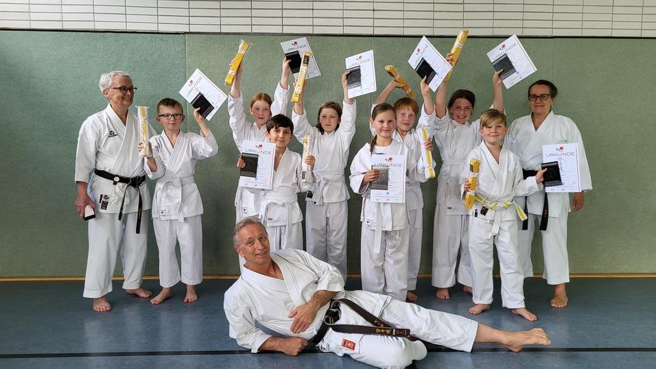 Gruppe von Karateka in Uniform hält Urkunden und Medaillen, während ein Trainer entspannt im Vordergrund posiert.