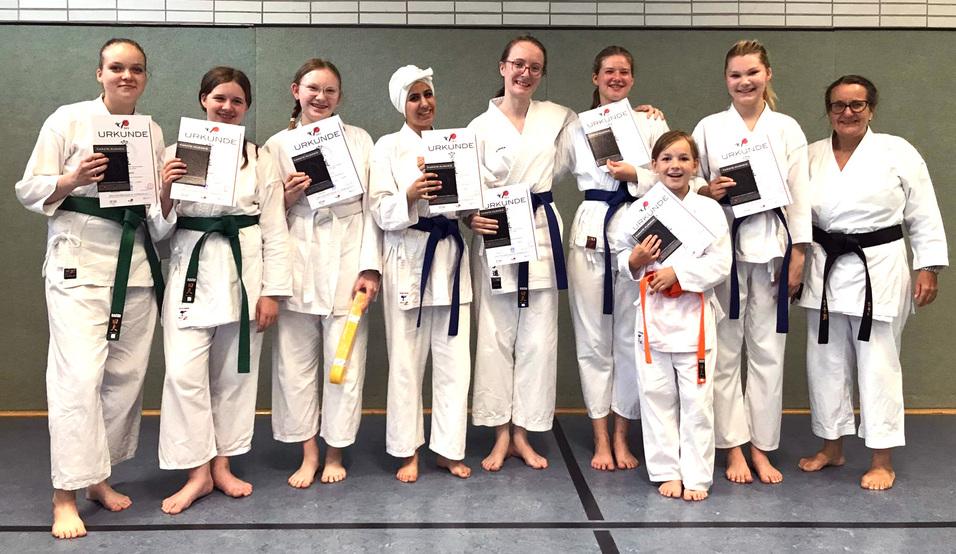 Gruppe von Karateka in weißen Gi mit Urkunden, die Erfolge feiern, stehen barfuß auf einer Matte.