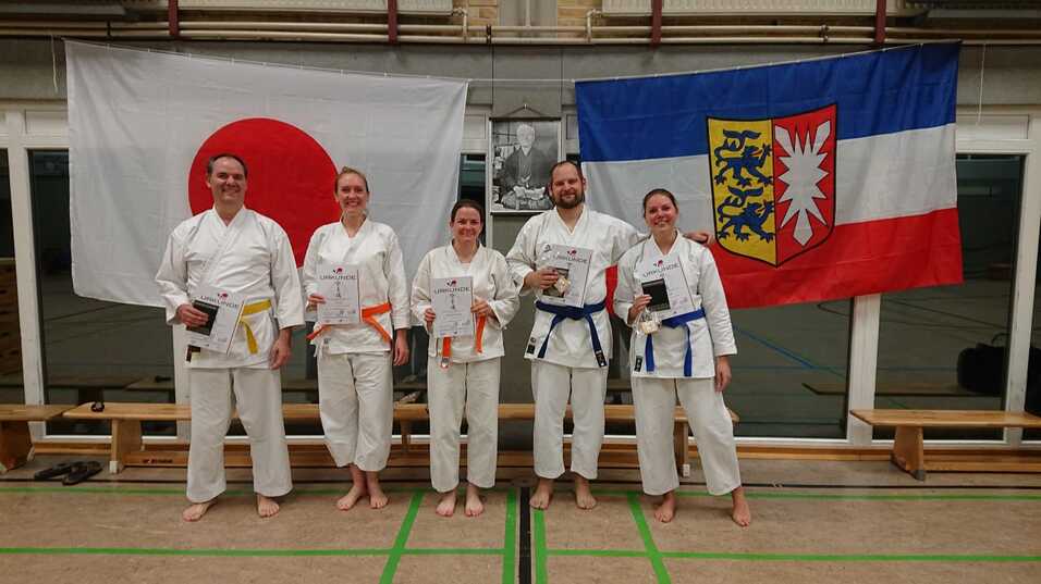 Fünf Karateka in weißen Anzügen halten Urkunden und stehen vor japanischen und deutschen Flaggen.