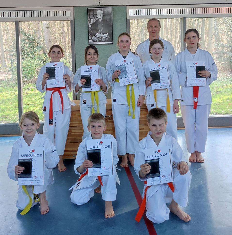 Eine Gruppe von Kindern in Karateanzügen hält Urkunden in der Hand, stehen vor einem Fenster mit Blick ins Freie.