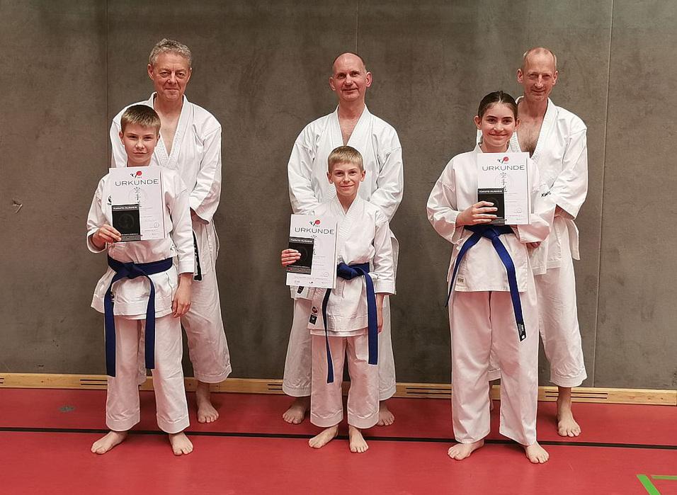 Gruppe von Karateka in weißen Gi mit blauen Gürteln, die stolz ihre Urkunden halten, auf einem roten Boden.