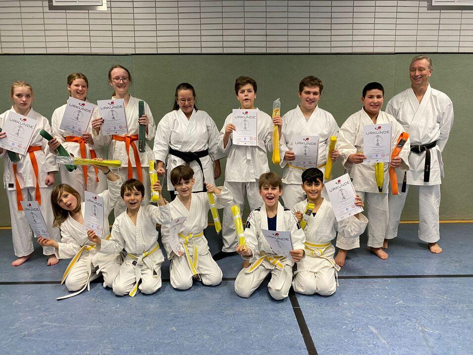 Gruppe junger Karateka in weißen Anzügen hält Urkunden und Gürtel in verschiedenen Farben auf einer Kampfmatte.