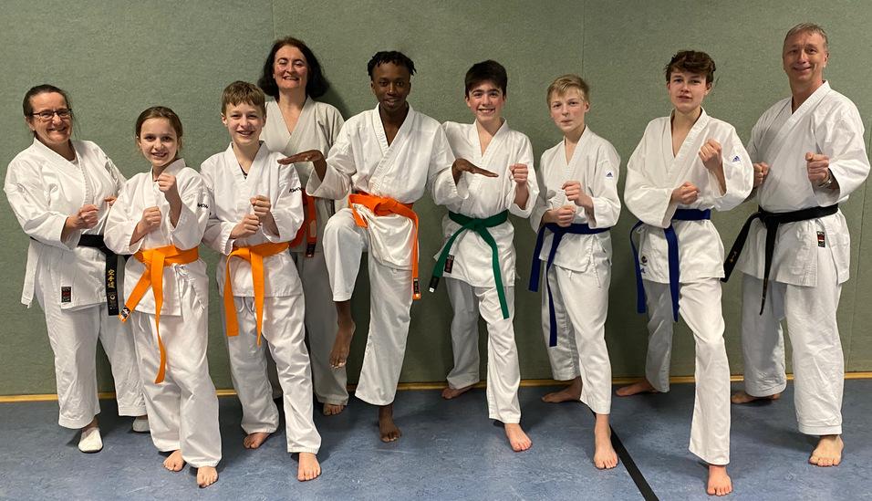 Gruppierung von acht Karateka in weißen Gi, mit verschiedenen Gürtelfarben, posierend in einem Trainingsraum.