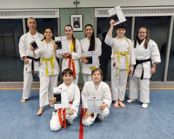 Gruppe von Karateka in weißen Anzügen hält Urkunden in der Sporthalle, einen Lehrer mit schwarzem Gürtel im Hintergrund.