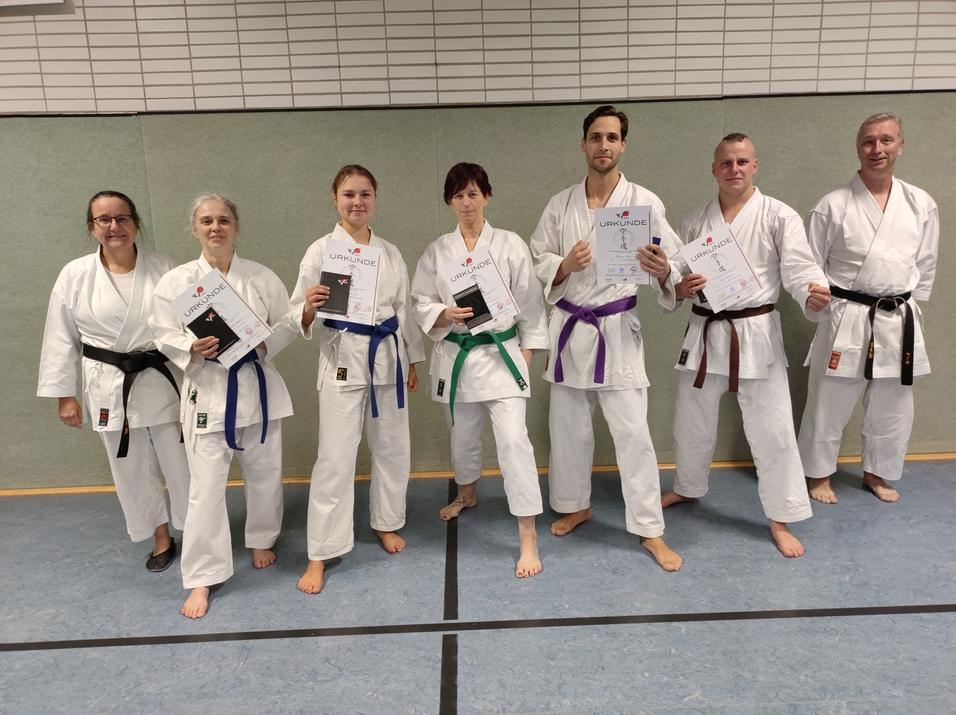 Gruppe von Karateka in weißem Gi hält Urkunden, stehen auf einer Matte in einer Sporthalle.
