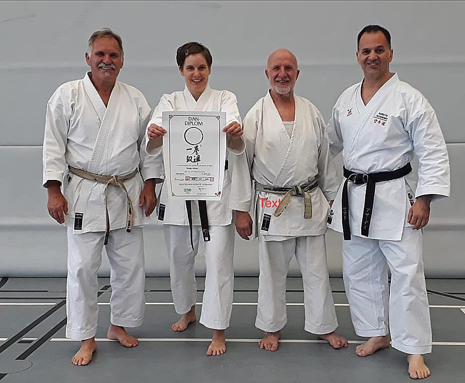 Vier Karateka in weißen Trainingsanzügen posieren gemeinsam mit einem Dan-Diplom auf einer Matte.