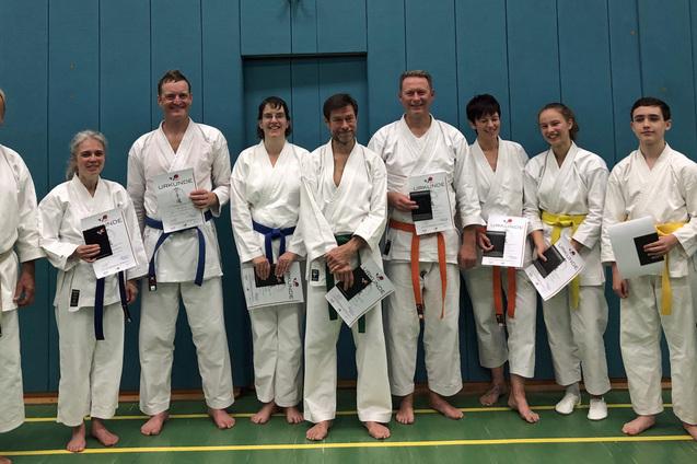 Gruppenfoto von Karatekas in wei&szlig;er Uniform, die stolz Urkunden in den H&auml;nden halten, vor einer blauen Wand.