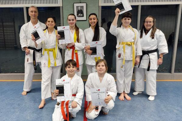 Gruppe von Karateka in wei&szlig;en Anz&uuml;gen h&auml;lt Urkunden in der Sporthalle, einen Lehrer mit schwarzem G&uuml;rtel im Hintergrund.