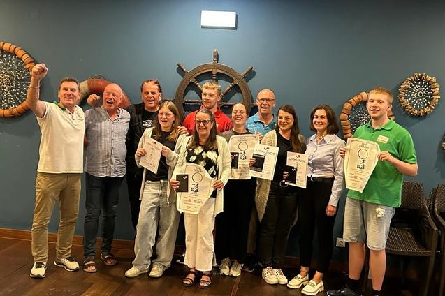 Gruppe von Personen mit Urkunden, l&auml;chelnd und feiernd, vor einer blauen Wand mit maritimen Dekorationen.