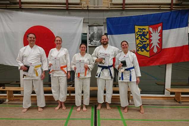 F&uuml;nf Karateka in wei&szlig;en Anz&uuml;gen halten Urkunden und stehen vor japanischen und deutschen Flaggen.