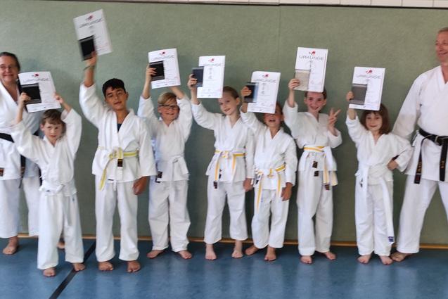 Gruppe junger Karateka in Gi h&auml;lt stolz Urkunden in einem Trainingsraum. Ein Trainer steht l&auml;chelnd daneben.