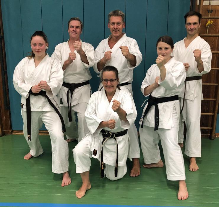 Sechs Karatekämpfer in weißen Anzügen und schwarzen Gürteln stehen grinsend in einer Kampfsporthalle.