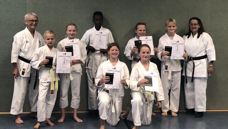 Gruppenfoto von Karateka in weißem Gi, die Urkunden in den Händen halten, im Hintergrund eine grüne Wand.