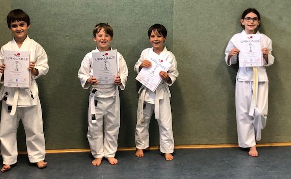 Vier Kinder in Judoanzügen halten Urkunden in der Hand, stehend vor einer grünen Wand in einem Sportraum.
