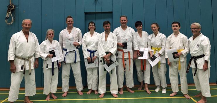 Gruppenfoto von Karatekas in weißer Uniform, die stolz Urkunden in den Händen halten, vor einer blauen Wand.