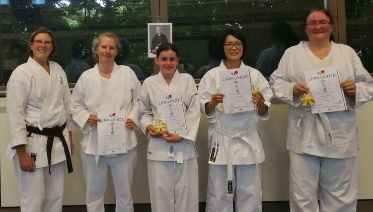 Fünf Karateka in weißen Gi halten Urkunden und Medaillen, lächeln vor einer Fensterfront mit einem Portrait im Hintergrund.