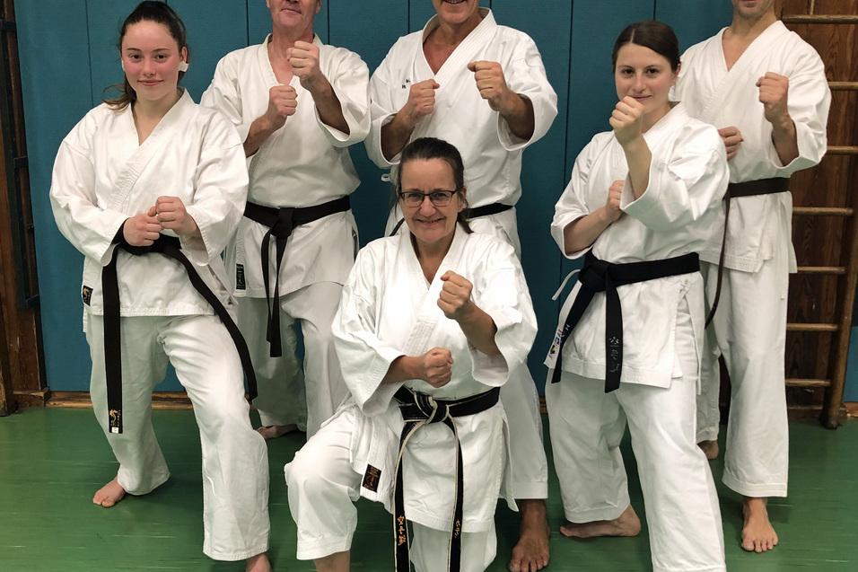 Sechs Karatek&auml;mpfer in wei&szlig;en Anz&uuml;gen und schwarzen G&uuml;rteln stehen grinsend in einer Kampfsporthalle.