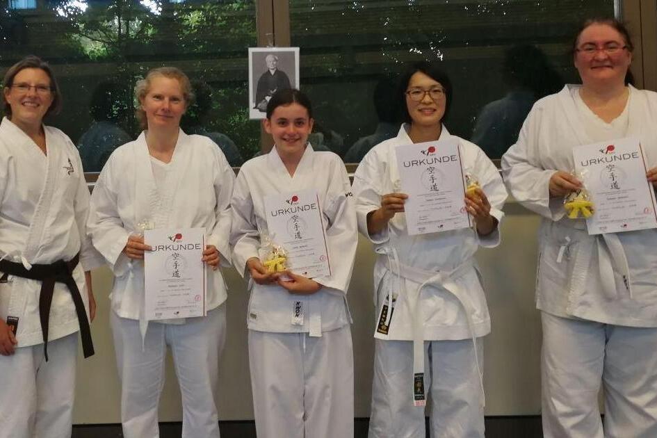 F&uuml;nf Karateka in wei&szlig;en Gi halten Urkunden und Medaillen, l&auml;cheln vor einer Fensterfront mit einem Portrait im Hintergrund.