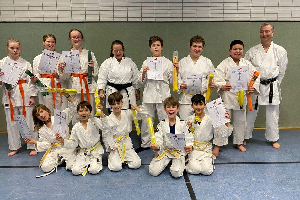 Prüfung 2022 Gruppe junger Karateka in weißen Anzügen hält Urkunden und Gürtel in verschiedenen Farben auf einer Kampfmatte.