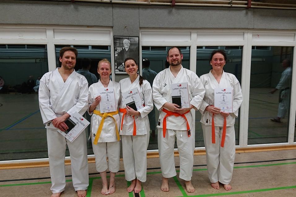 F&uuml;nf Karateka in wei&szlig;en Gi mit Urkunden stehen l&auml;chelnd vor einem Fenster in einem Dojo.