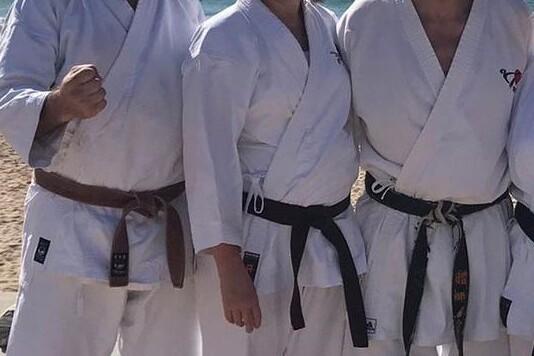 Drei Karateka in wei&szlig;en Gi mit Braungurt und zwei Schwarzgurten posieren l&auml;chelnd am Strand vor dem Meer.