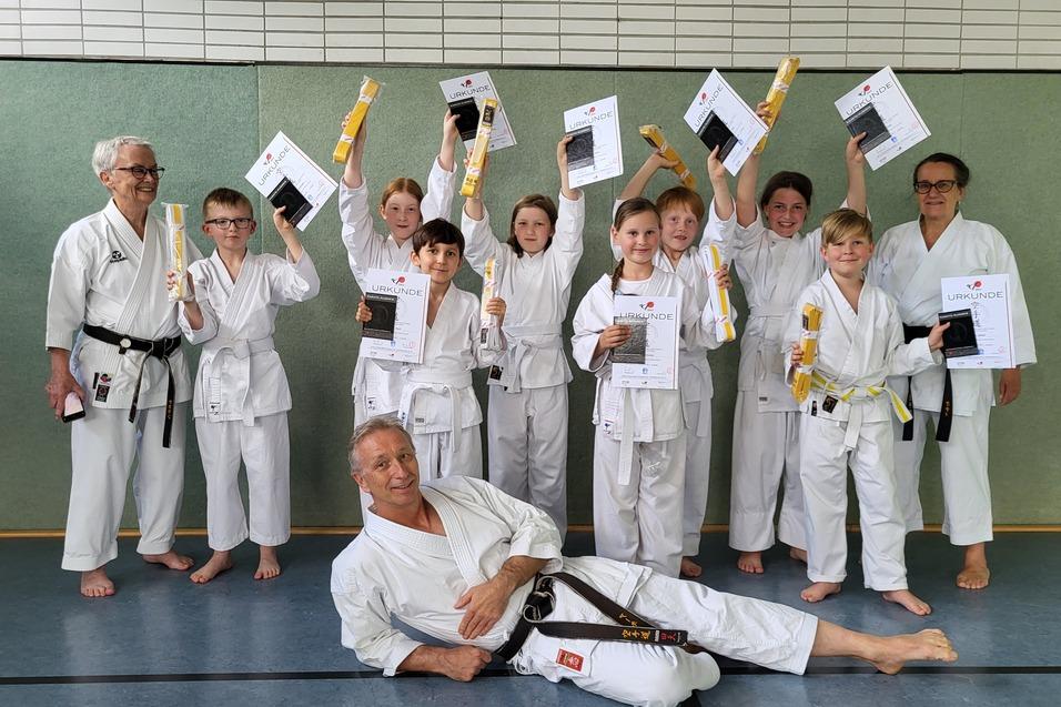 Gruppe von Karateka in Uniform h&auml;lt Urkunden und Medaillen, w&auml;hrend ein Trainer entspannt im Vordergrund posiert.