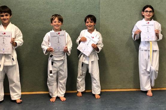 Vier Kinder in Judoanz&uuml;gen halten Urkunden in der Hand, stehend vor einer gr&uuml;nen Wand in einem Sportraum.