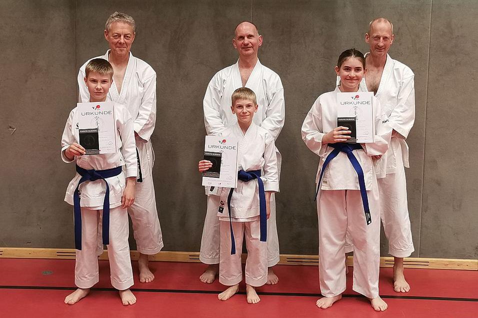 Gruppe von Karateka in wei&szlig;en Gi mit blauen G&uuml;rteln, die stolz ihre Urkunden halten, auf einem roten Boden.